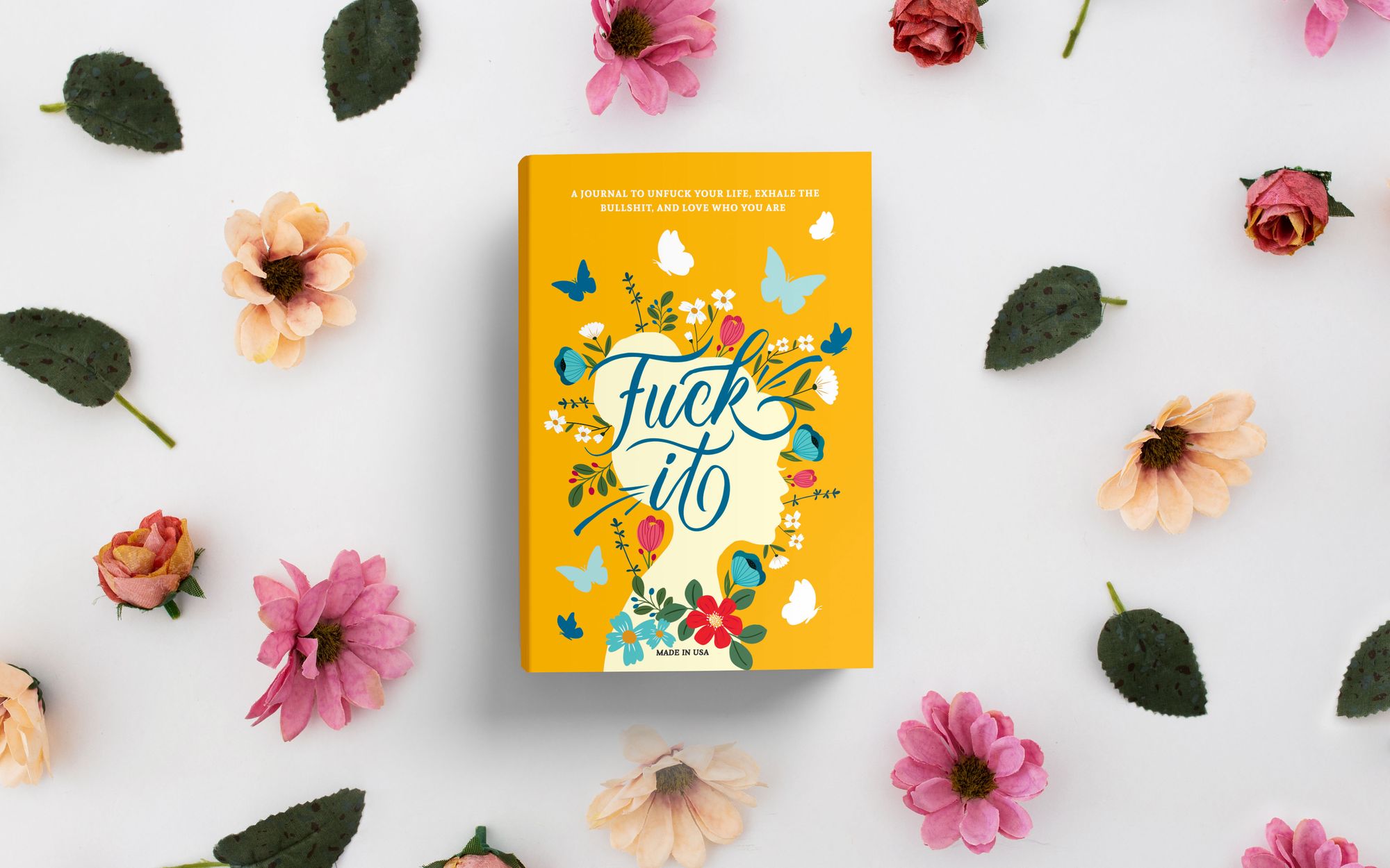 Fuck It journal, yellow cover, among pink flowers and leaves